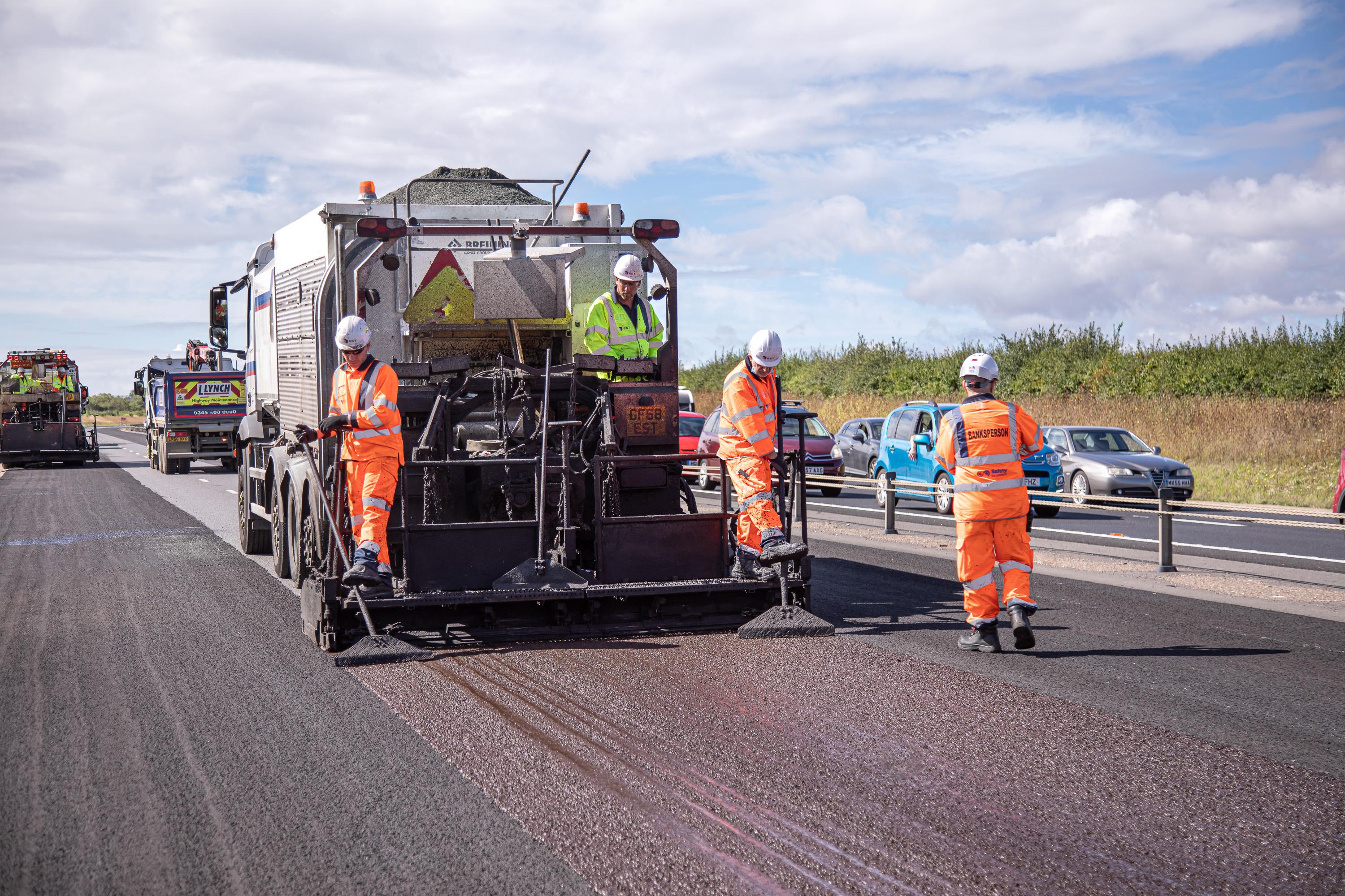 Highway workers in high-visibility clothing laying specialist warm mix asphalt on a road surface using machinery, supporting carbon reduction through lower-temperature asphalt production.
